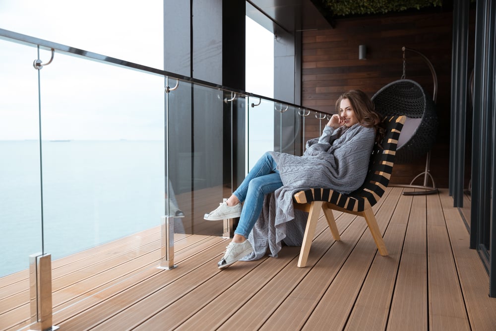 Smiling young woman sitting wrapped in knitted coverlet on the glass balcony